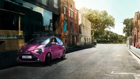 Pink Toyota Aygo driving on an empty city street lined with modern and historic buildings under bright daylight.