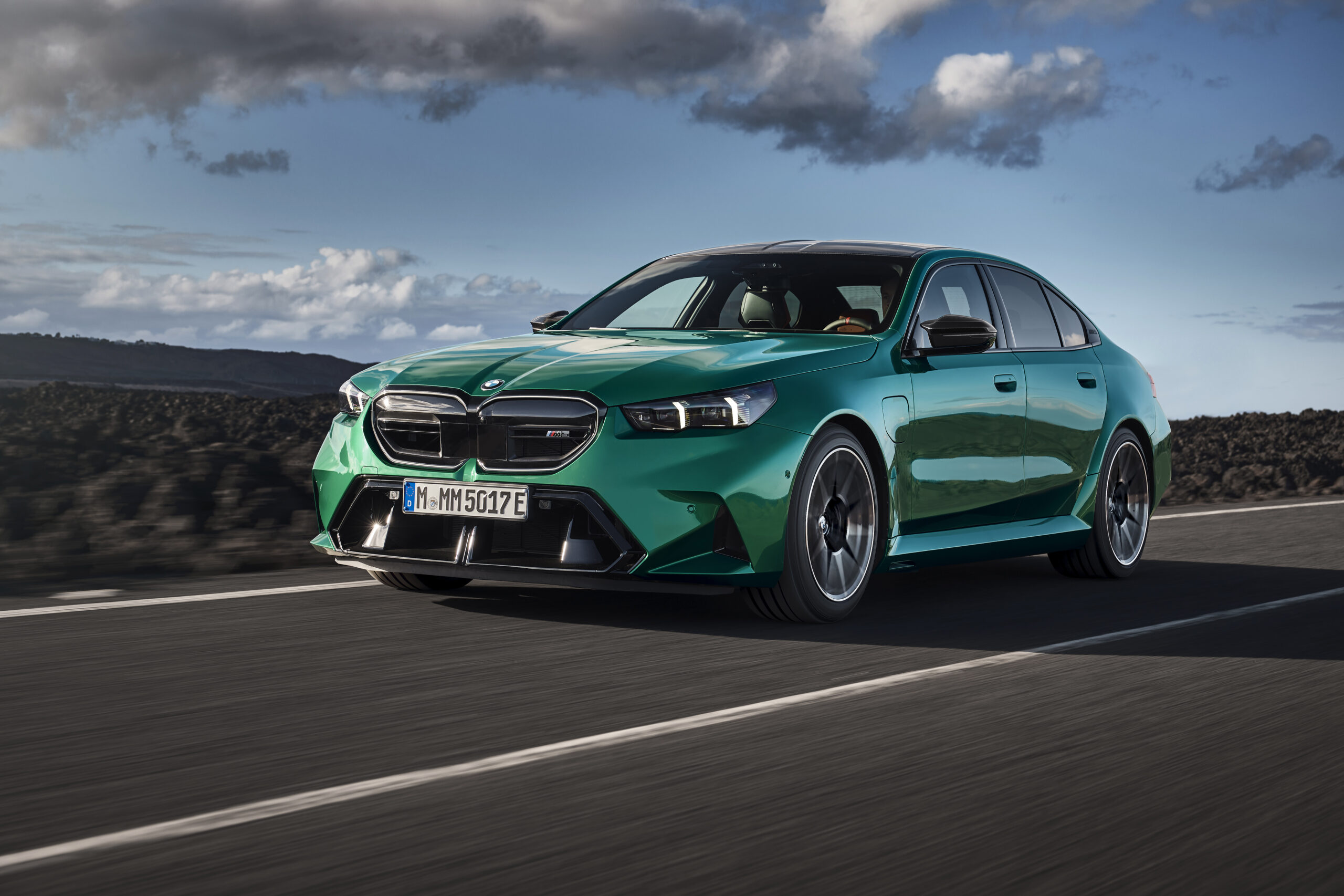 Green BMW M5 speeding on an open road with mountains in the background, showcasing its sleek design and powerful stance.
