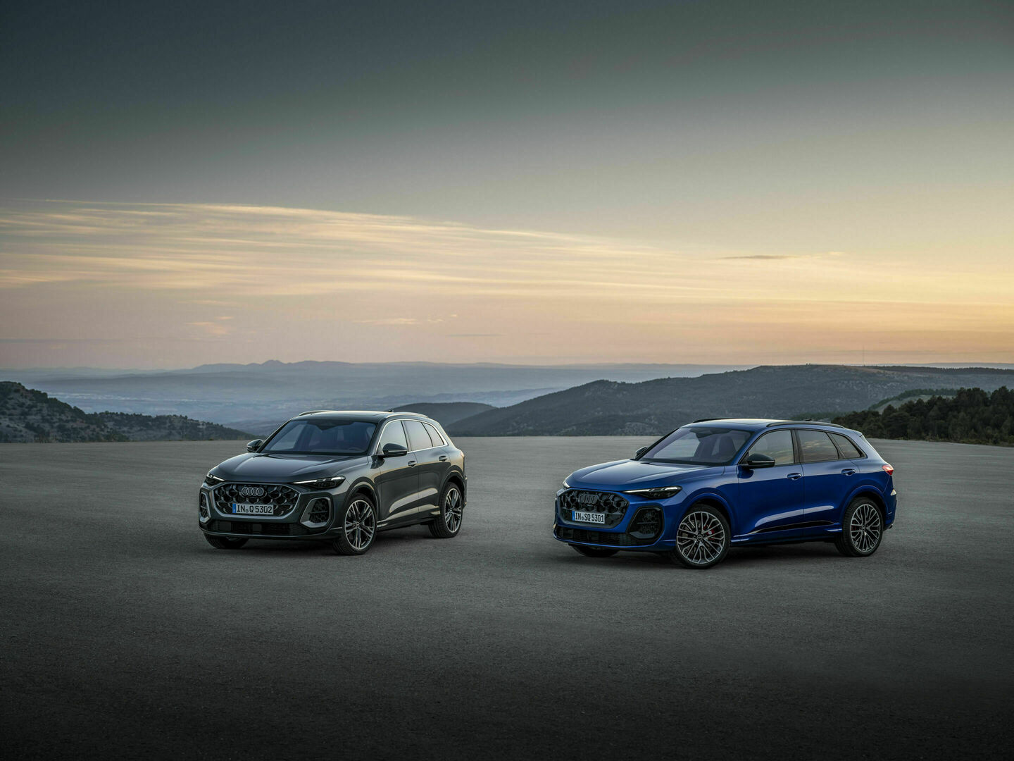 Audi Q5 in black and Audi SQ5 TFSI in blue parked side by side on a vast open road at sunset, highlighting their sleek designs.