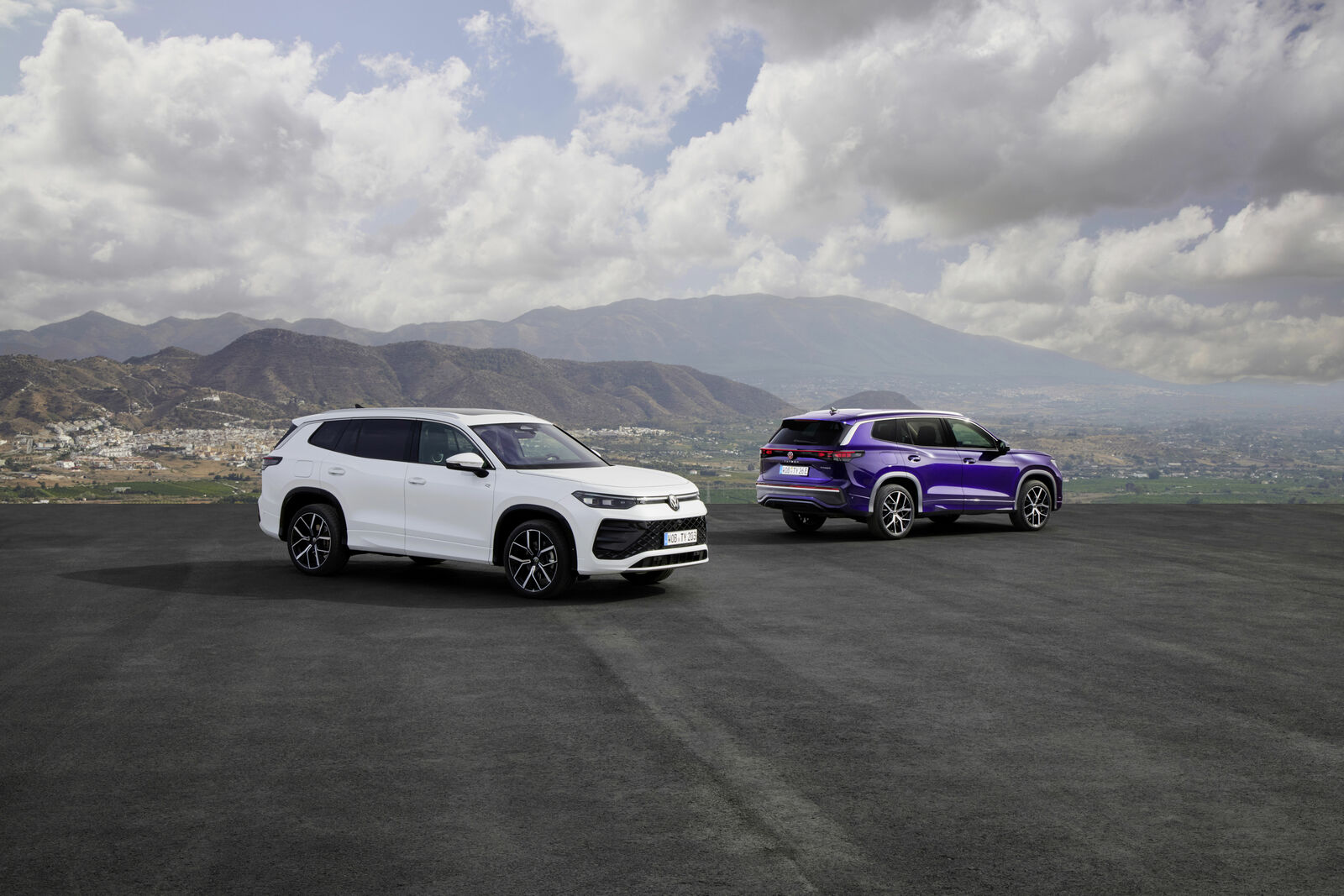 White and purple Volkswagen Tayron SUVs parked on a wide road with a mountainous landscape and cloudy sky in the background.