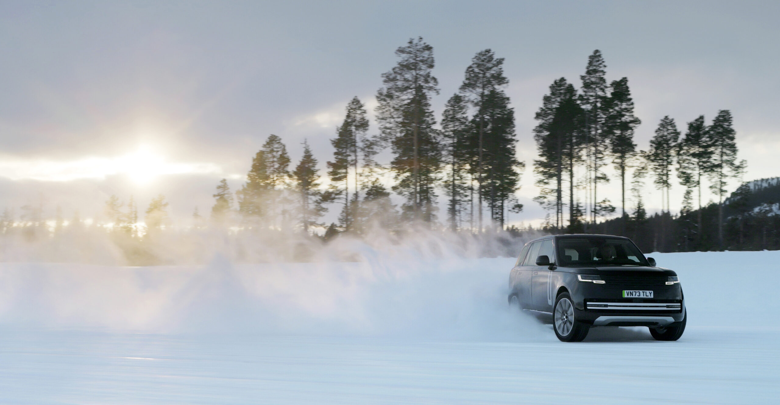 Range Rover Electric SUV driving through snow with a cloud of powder behind it, set against a backdrop of trees and a winter sunset.
