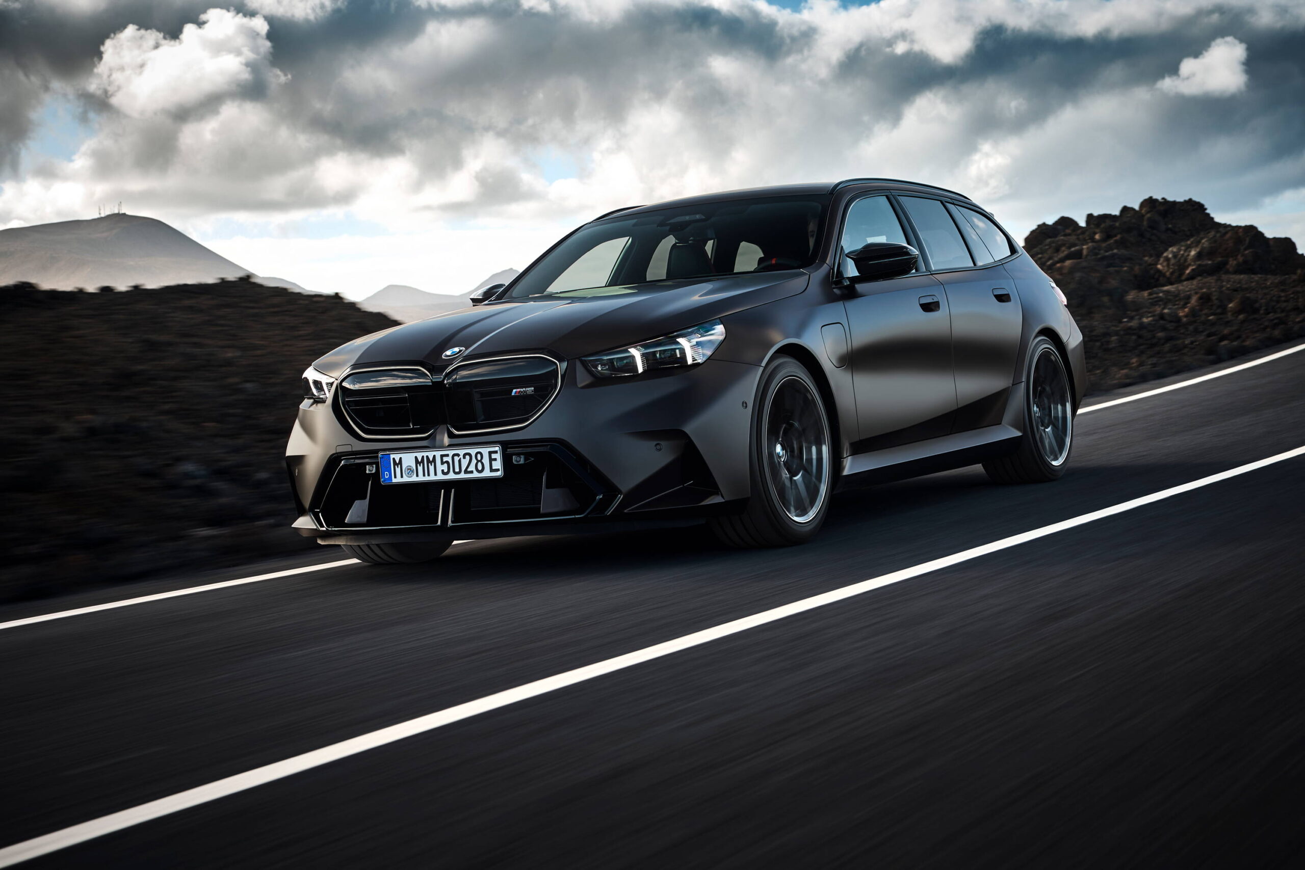 Sleek BMW M5 Touring driving on a scenic highway with a mountainous backdrop under cloudy skies.