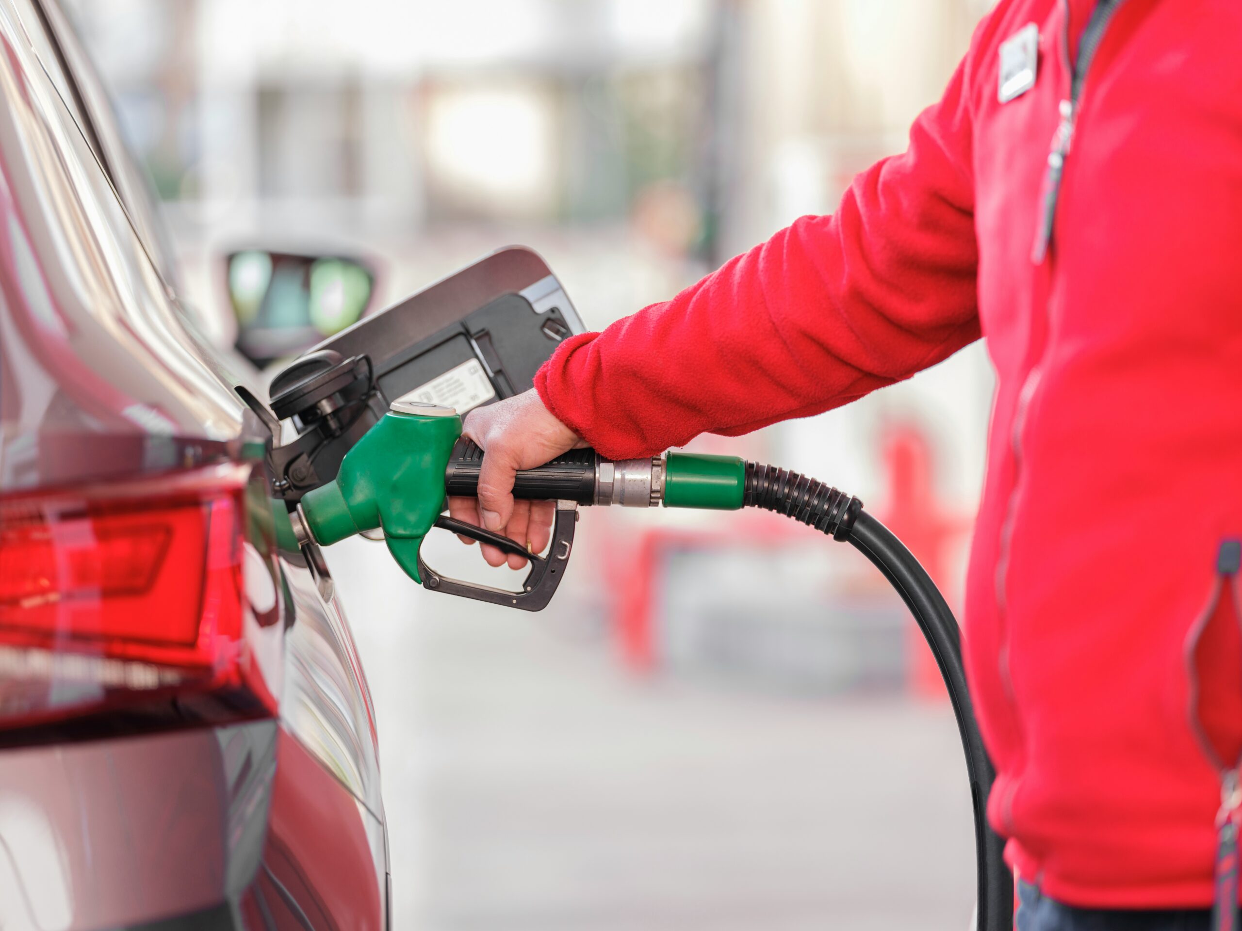 Attendant in red jacket refueling a car with a green gas nozzle at a station, showing the rear side of the vehicle.