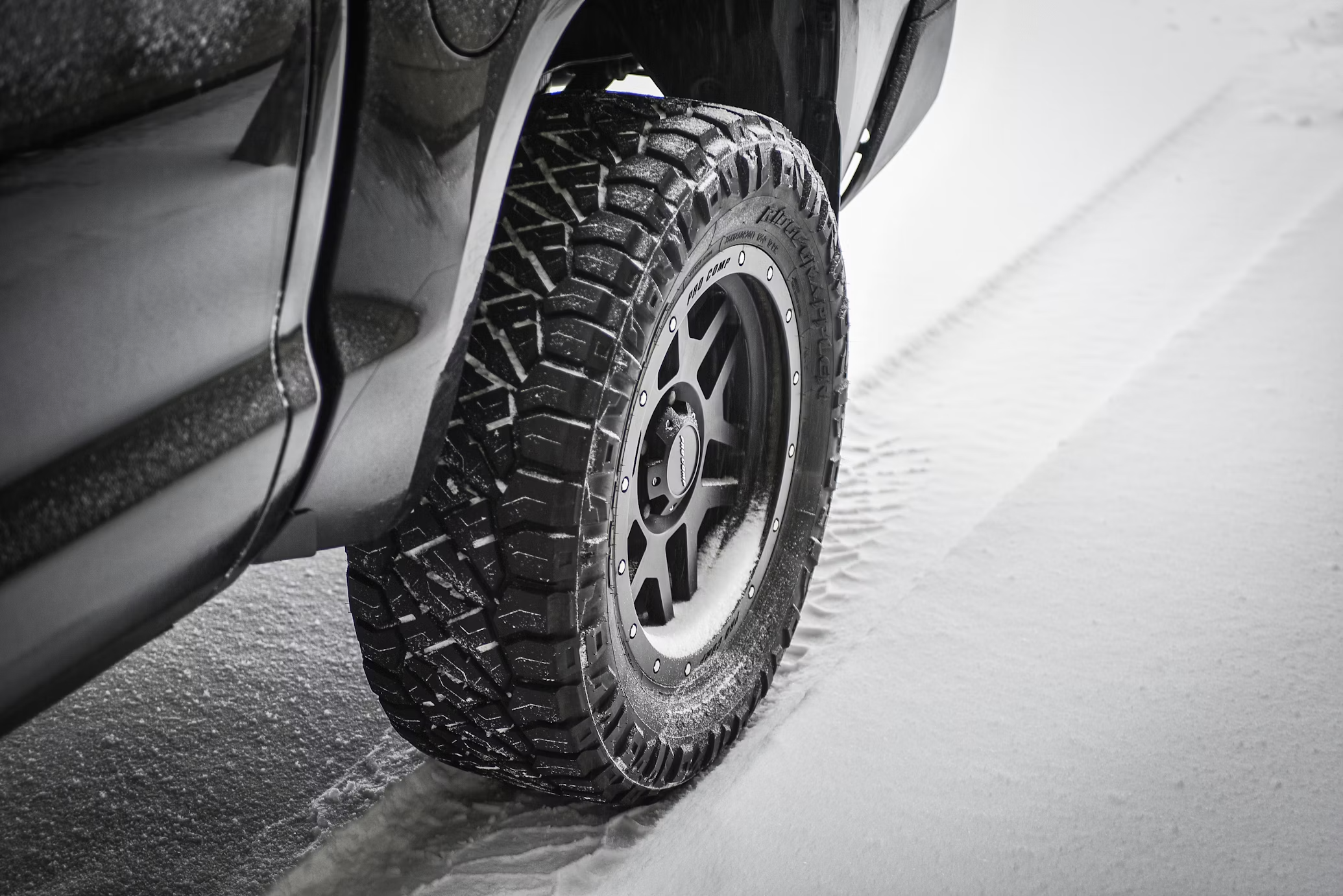 Off-road tire with deep treads on a black vehicle driving through snow, showcasing traction on snowy terrain.