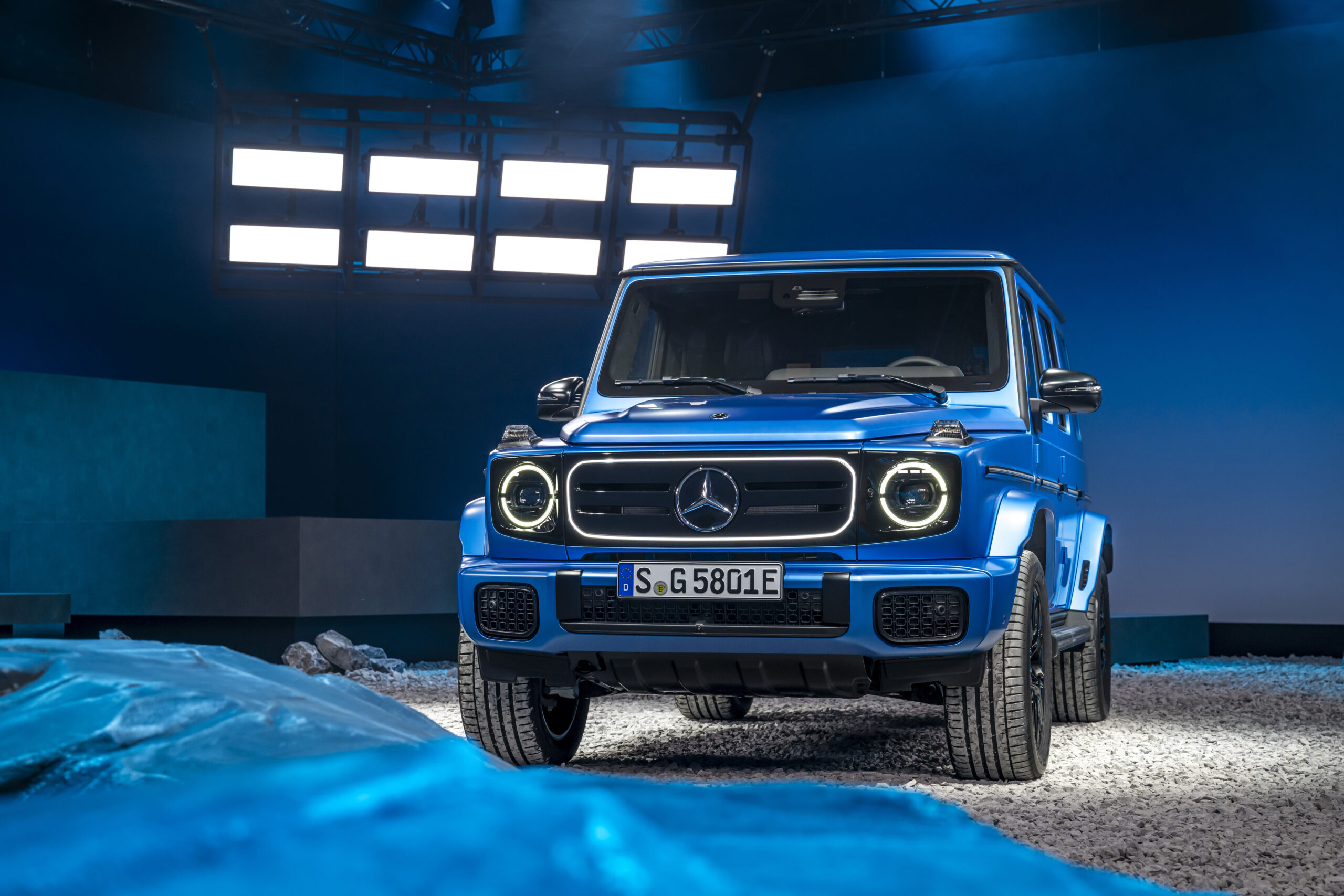 Front view of a blue Mercedes-Benz G 580 with EQ Technology, featuring circular LED headlights and a bold grille in a studio setting.