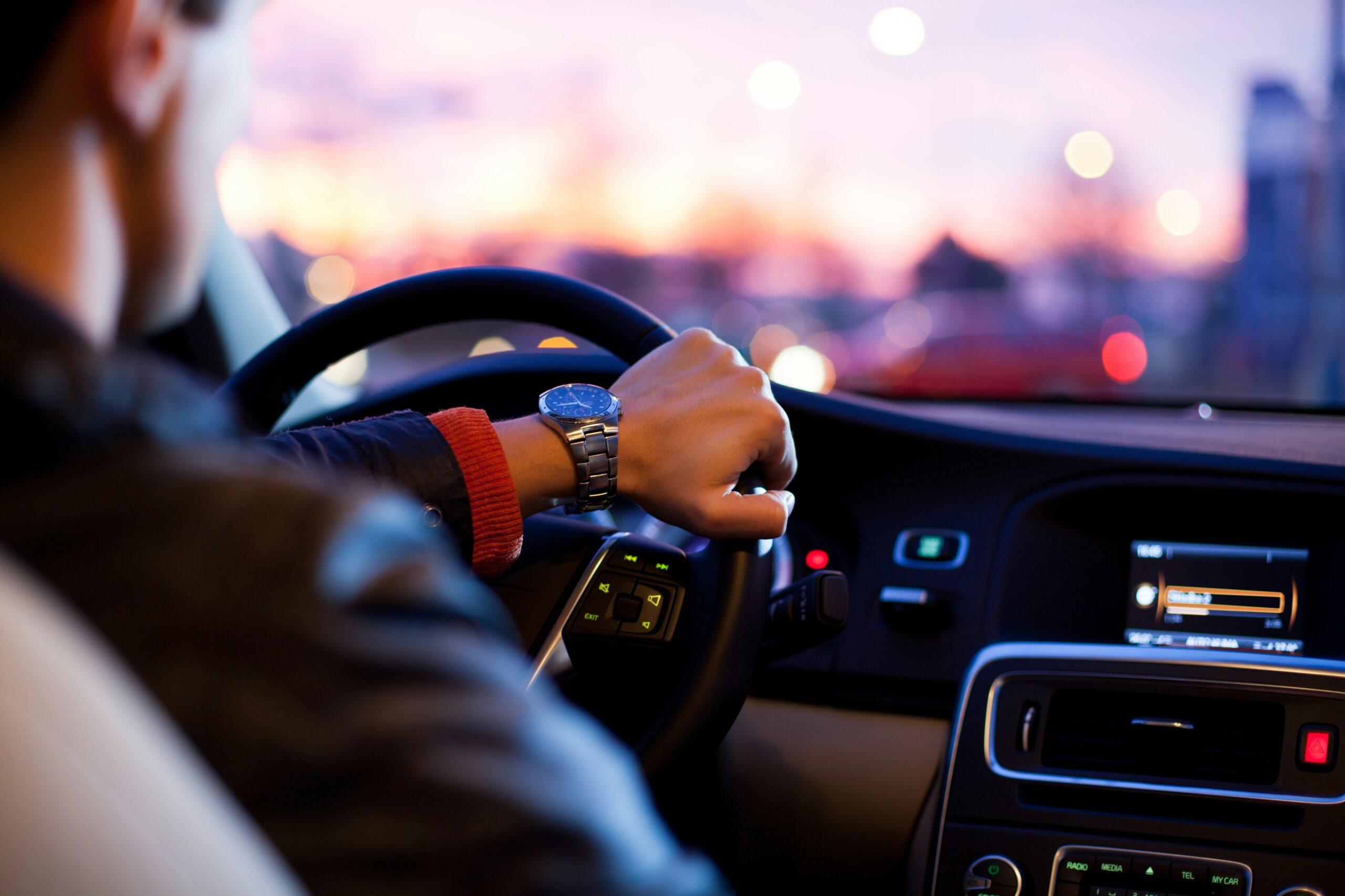 Driver navigating a car equipped with advanced safety features, with a view of the dashboard and steering wheel controls at sunset.