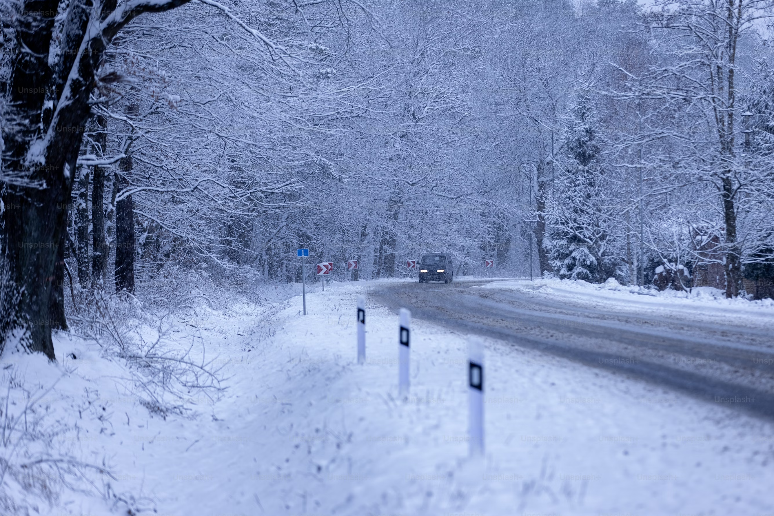 Car driving on a snow-covered forest road, surrounded by snow-laden trees and road signs, depicting winter conditions.